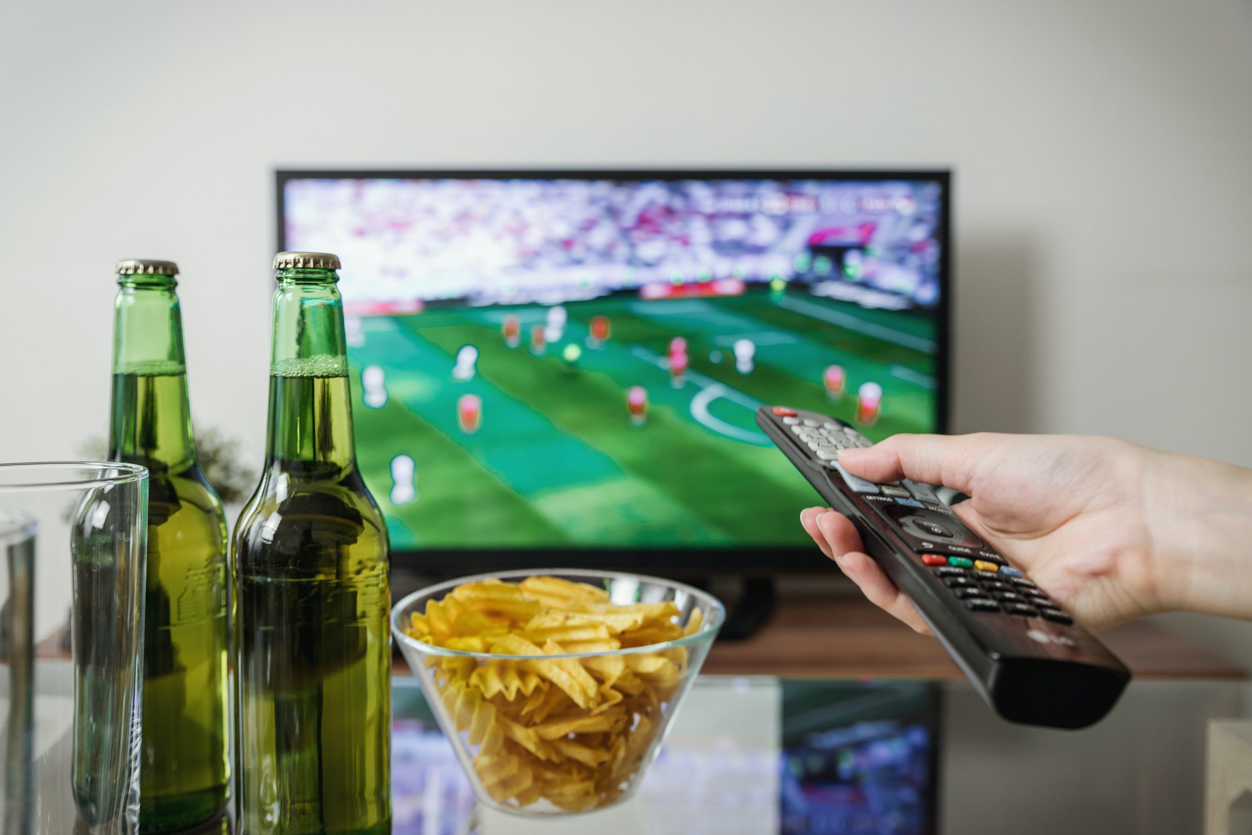 En primer plano, una mesa de cristal sobre la que hay dos cervezas y un bol de patatas. A la derecha, una mano con un mando de la televisión cambia de canal en el dispositivo que se ve al fondo, borroso.